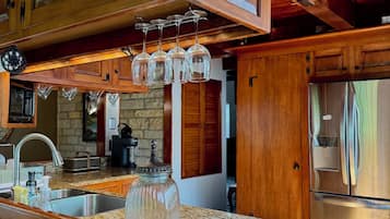 Une cuisine dotée de meubles en bois, de plans de travail en granit, d’un réfrigérateur en acier inoxydable et de verres à vin suspendus.