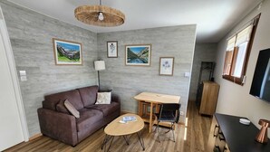 Uma sala de estar moderna com um sofá, mesa de centro e área de jantar com mesa e cadeiras. Há quadros nas paredes e uma TV fixada na parede.