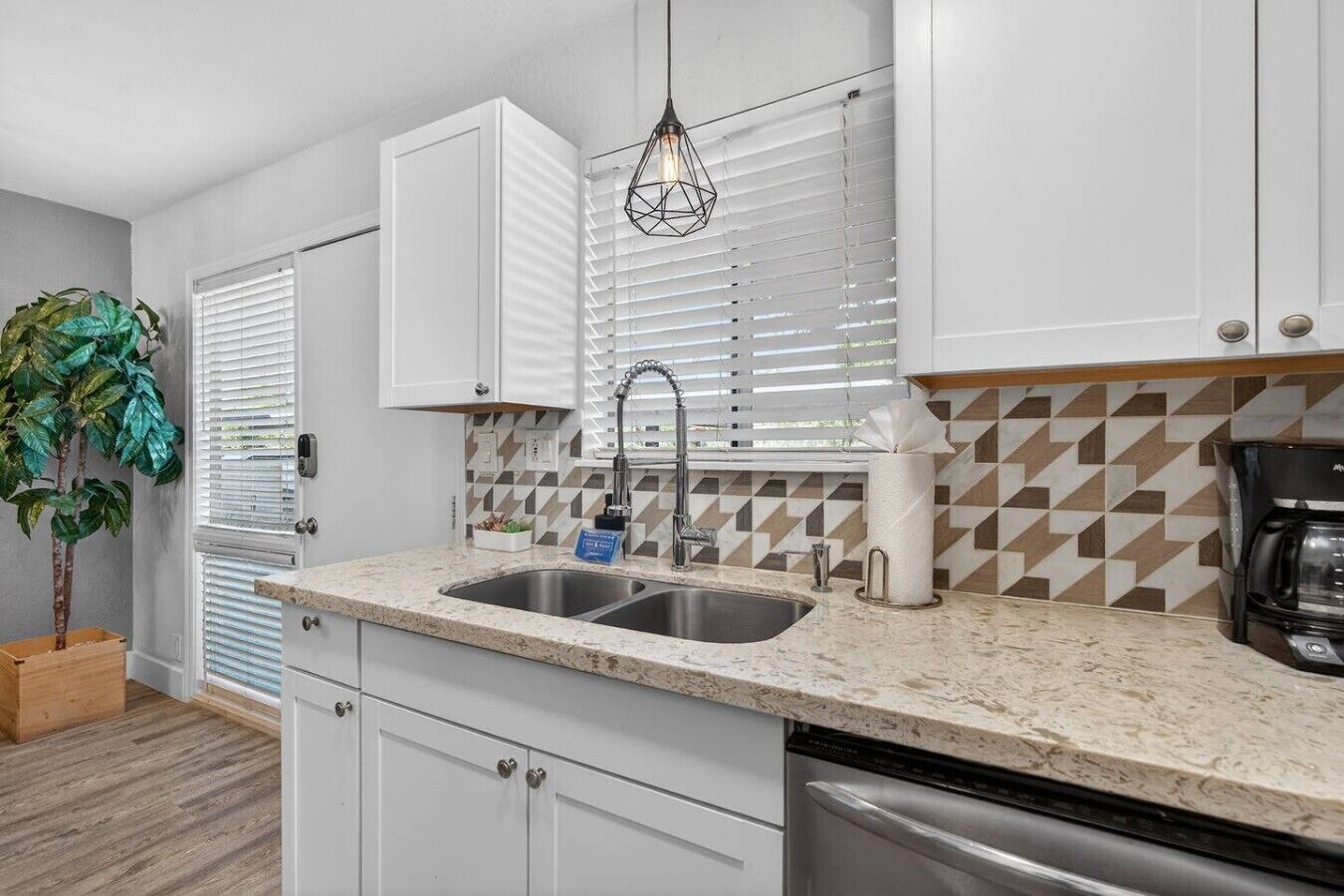 A modern kitchen with white cabinets, a granite countertop, a stainless steel sink, a coffee maker, and a potted plant.