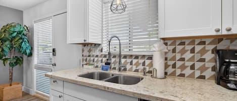 A modern kitchen with white cabinets, a granite countertop, a stainless steel sink, a coffee maker, and a potted plant.