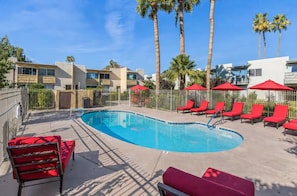 A swimming pool with red lounge chairs and umbrellas, surrounded by palm trees and a building. - Walk to Fashion Square | Heated Pool & EV Charger (Scottsdale)