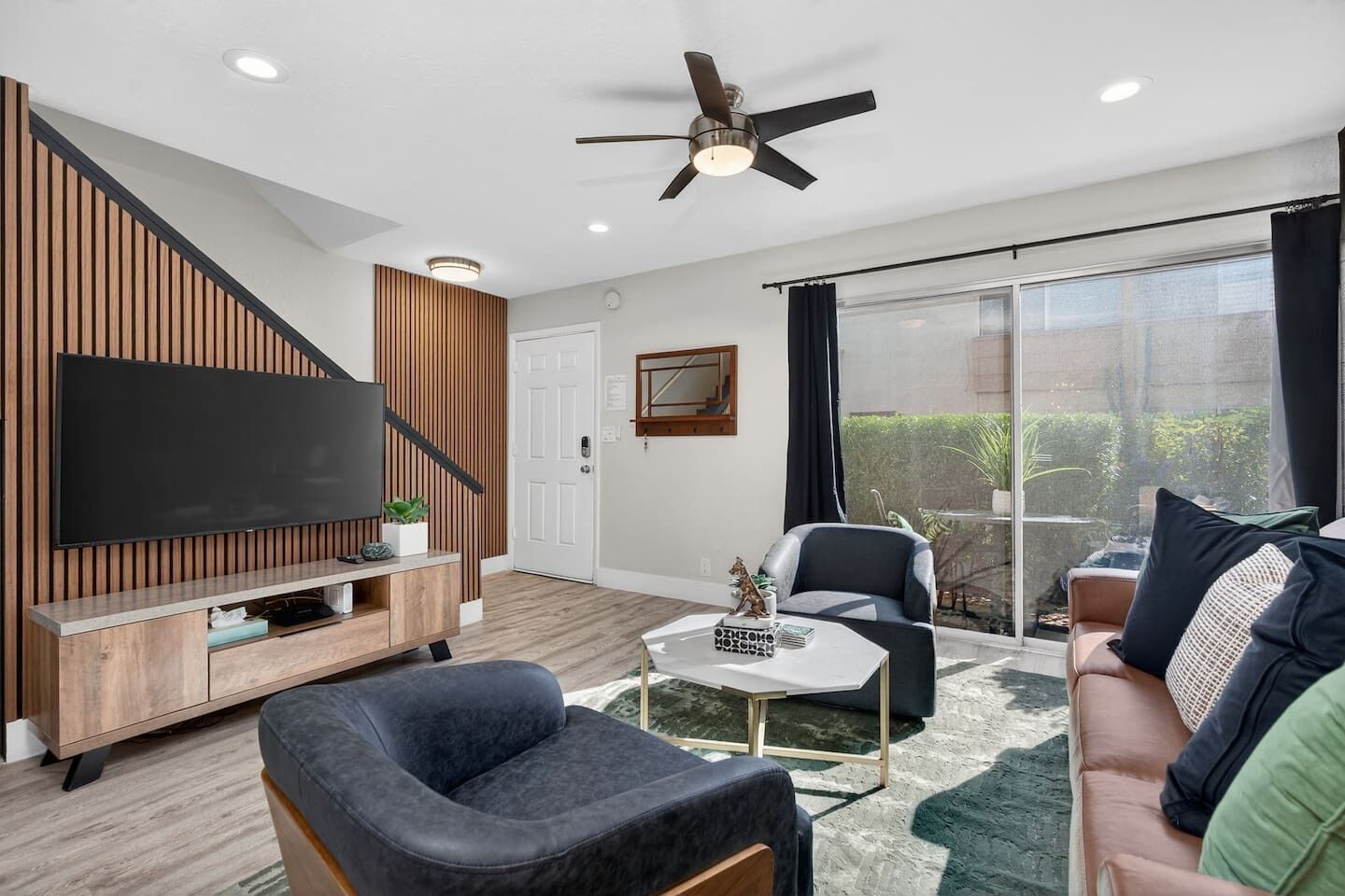 A modern living room with a flat-screen TV, a ceiling fan, a staircase, and a sliding glass door leading to a balcony.