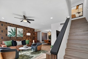 A modern living room with a brown brick wall, a ceiling fan, a staircase with a wooden finish, and a dining area in the background. - Walk to Fashion Square | Heated Pool & EV Charger (Scottsdale)