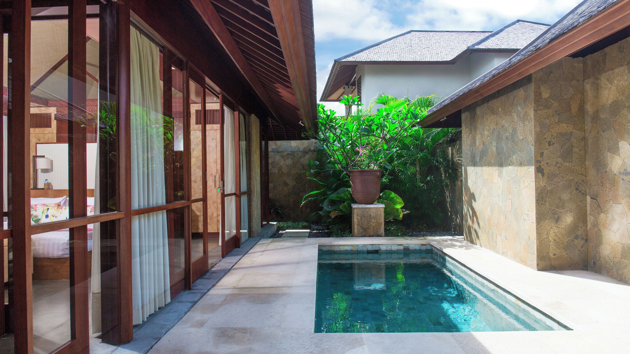 A pool area with a stone wall, a large potted plant, and a bed visible through a glass door.