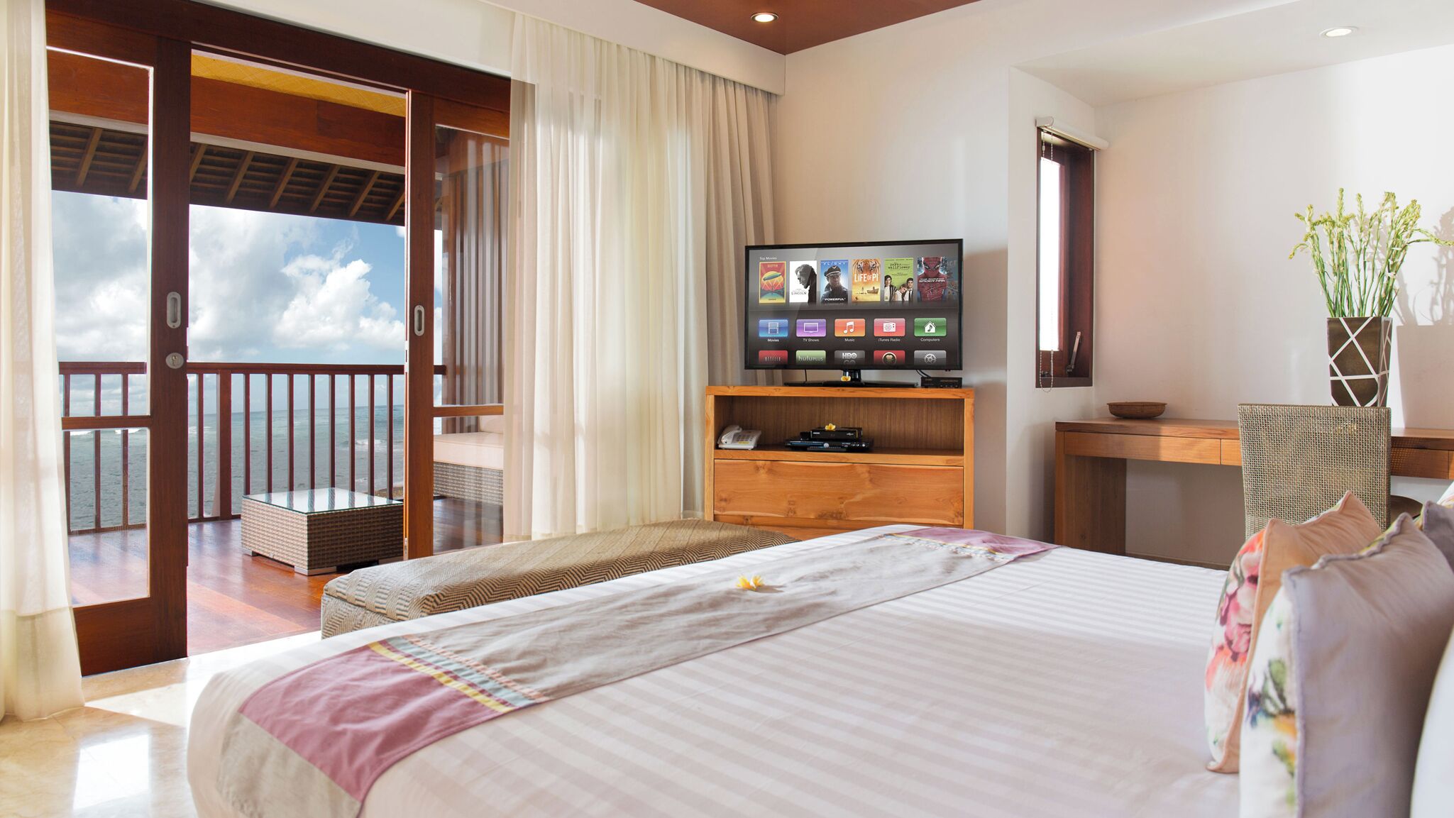 A hotel room with a large bed, a TV on a wooden stand, a desk, and a view of the ocean through a sliding door.