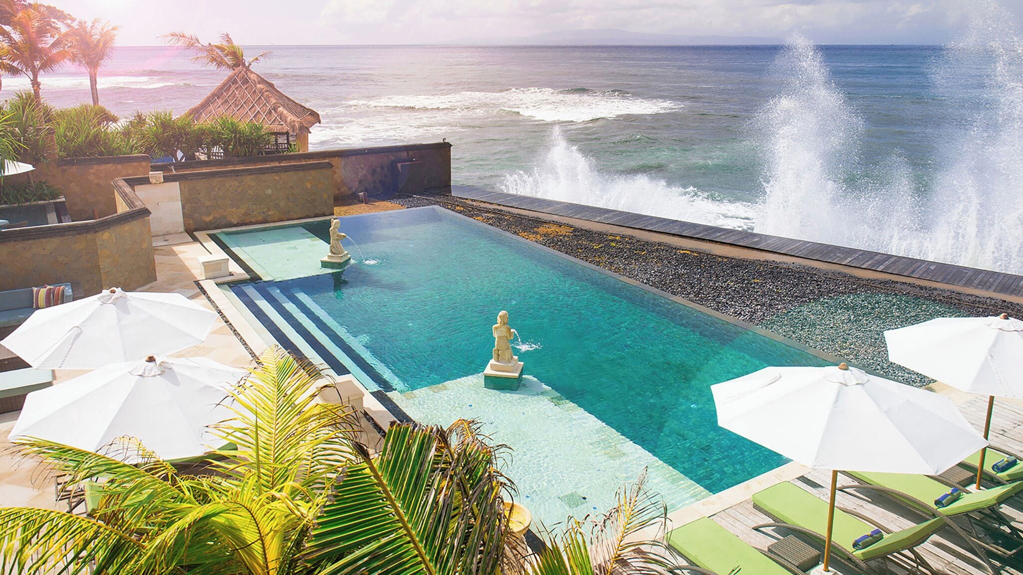 A pool with a statue, surrounded by lounge chairs and umbrellas, overlooking the ocean.