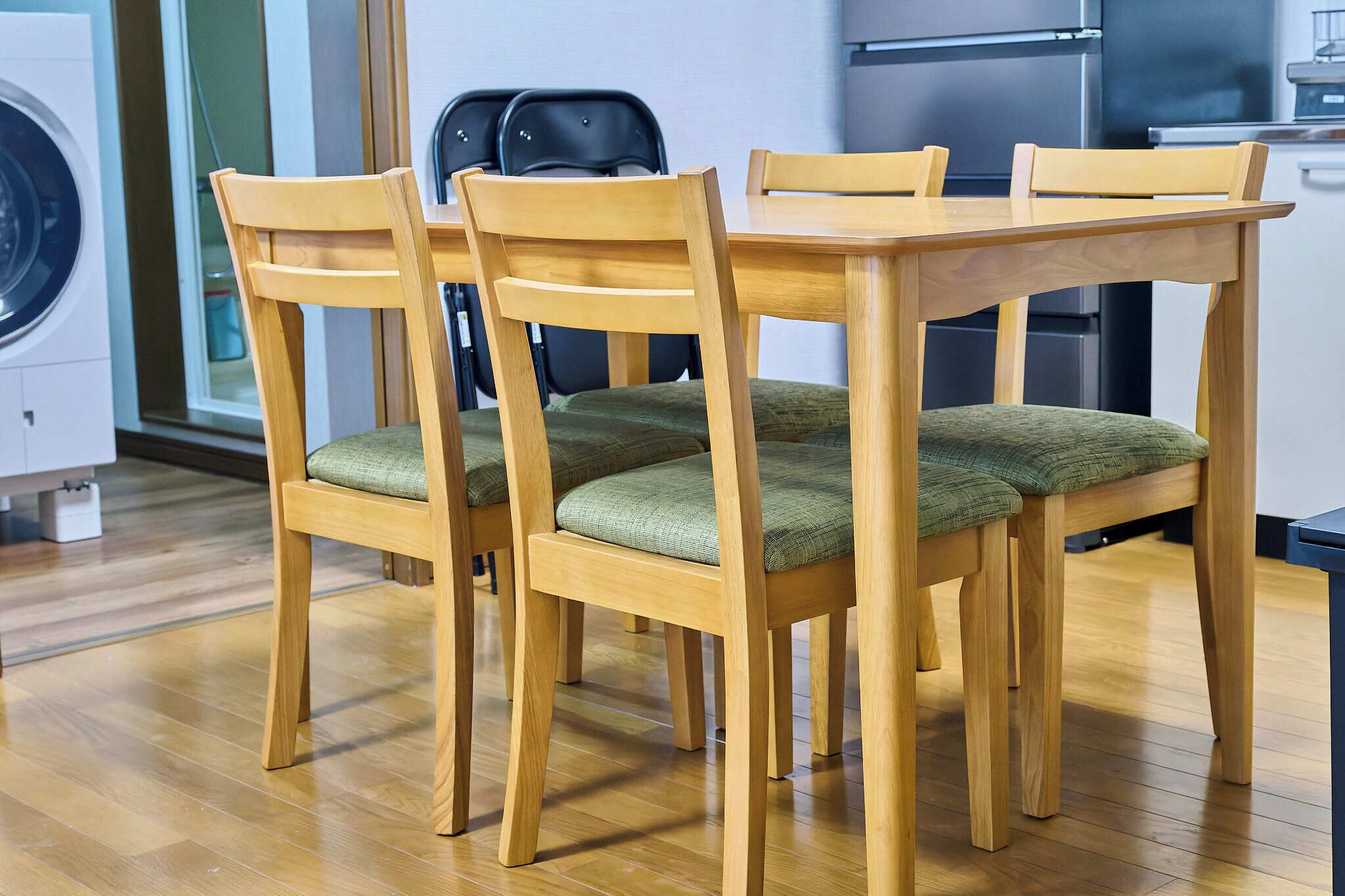 A dining area with wooden chairs and a table, a washing machine, and a kitchen area in the background.
