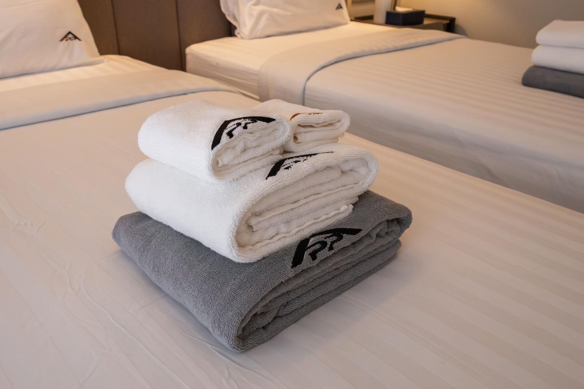Una cama bien hecha con ropa blanca y gris, una toalla doblada y una almohada con un logotipo.