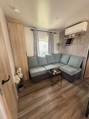 A modern living room with a sectional sofa, a small table, a potted plant, and a 'HOME' sign on the wall. - A Stay in Cap Ferret • Modern Mobile Home in Les Viviers (Lège-Cap-Ferret)