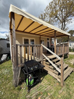 A covered outdoor area with a grill, wooden deck, and a vehicle in the background. - A Stay in Cap Ferret • Modern Mobile Home in Les Viviers (Lège-Cap-Ferret)
