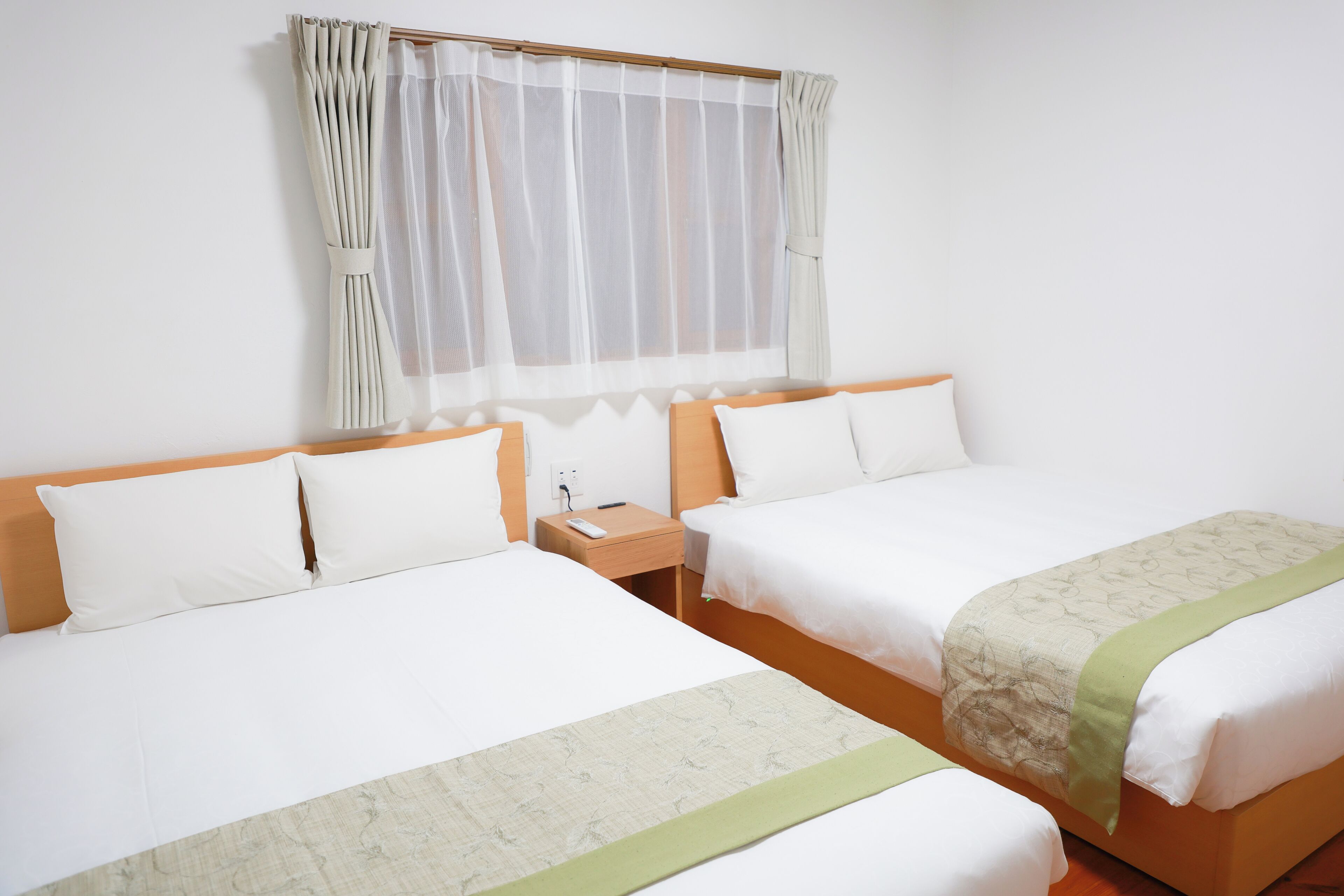 A hotel room with two beds, a nightstand, a window with sheer curtains, and a bedspread with a green accent.