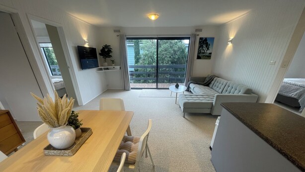 Un salon moderne comprenant un canapé, une table à manger avec des chaises, un téléviseur fixé au mur et un balcon offrant une vue sur les arbres.