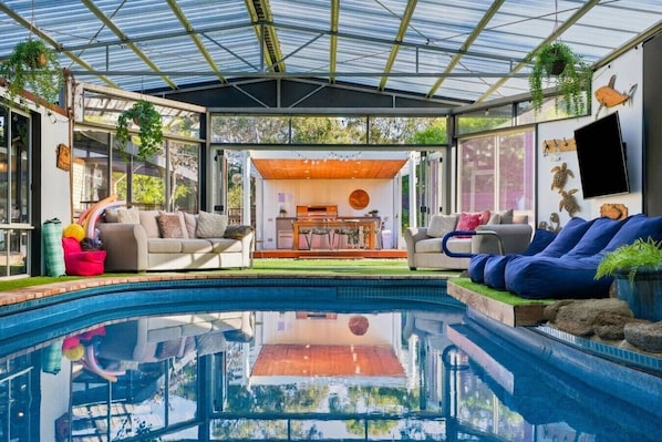 Uma área interna de piscina moderna com teto de vidro, dois sofás, uma mesa de jantar e uma televisão.