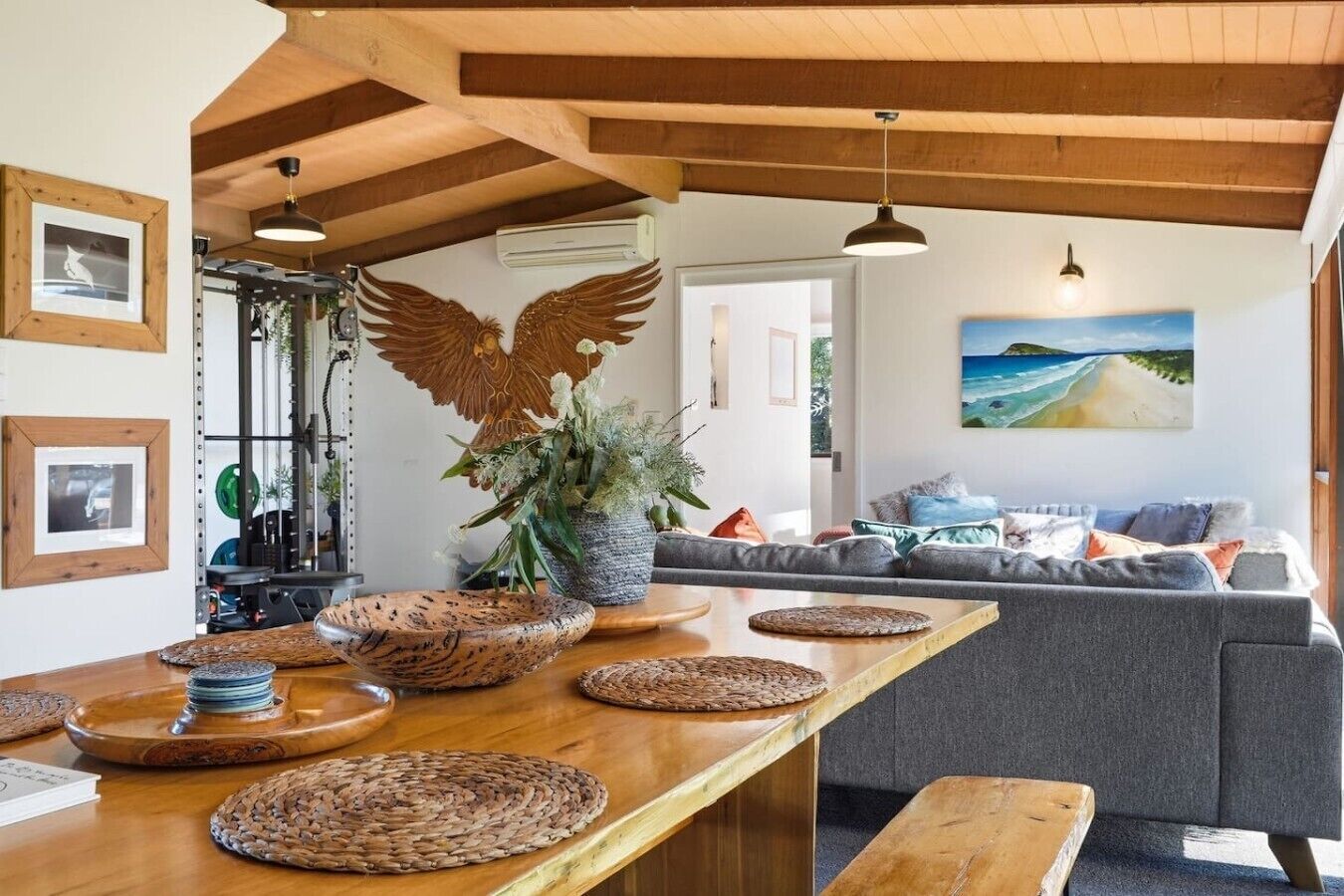 A modern living space with a wooden ceiling, a large wooden table with woven placemats, a grey sofa, and a wall-mounted air conditioner.