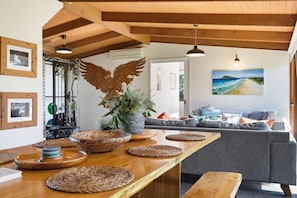Um espaço de estar moderno com teto de madeira, uma mesa de madeira grande com jogos americanos trançados, um sofá cinza e um ar condicionado fixado na parede.
