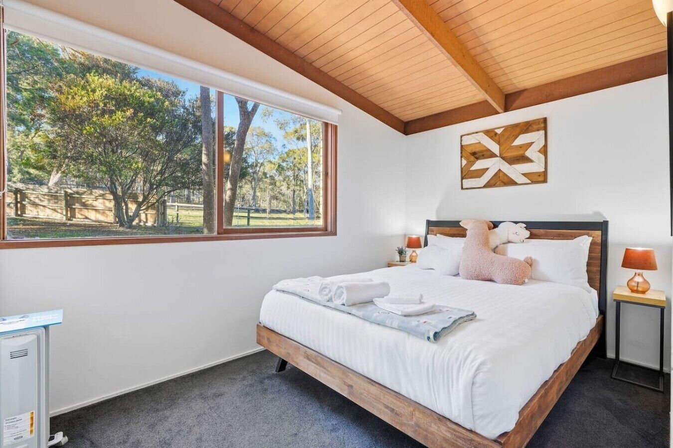 A bedroom with a large bed, a view of trees, a wooden ceiling, and a wall-mounted artwork.