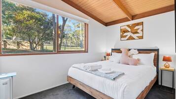 A bedroom with a large bed, a view of trees, a wooden ceiling, and a wall-mounted artwork.