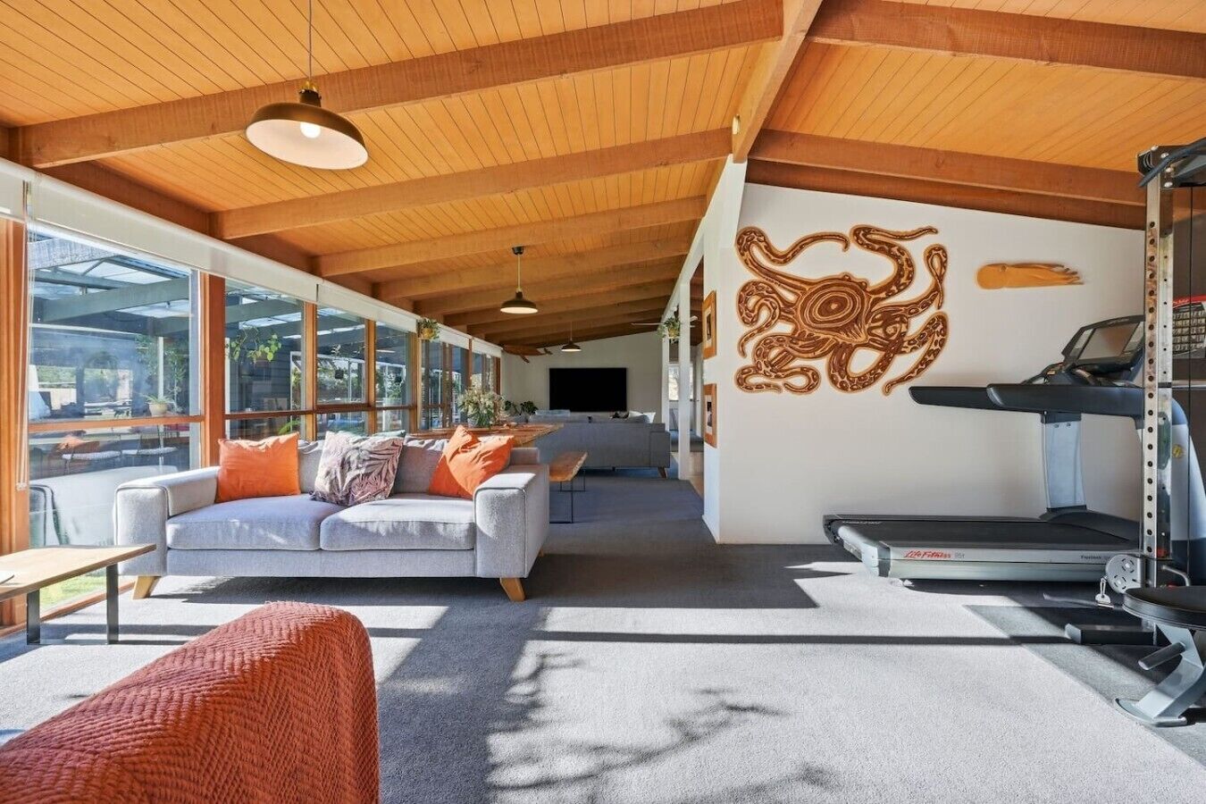 A modern living room with a grey sofa, orange cushions, a treadmill, and a large window.