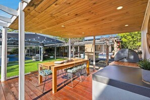 A covered outdoor dining area with a wooden table and chairs, a grill, and a view of a pool and garden. - Rural Getaway: Alpacas, Pool, Hot tub, Gym, Tennis (Sandford)