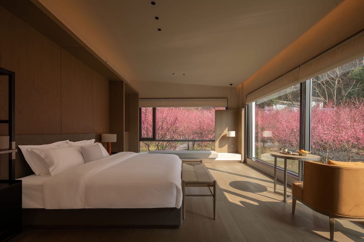 Une chambre moderne avec un grand lit, des tables de chevet, un banc, une chaise et une vue sur des arbres aux feuilles roses, visible à travers de grandes fenêtres.