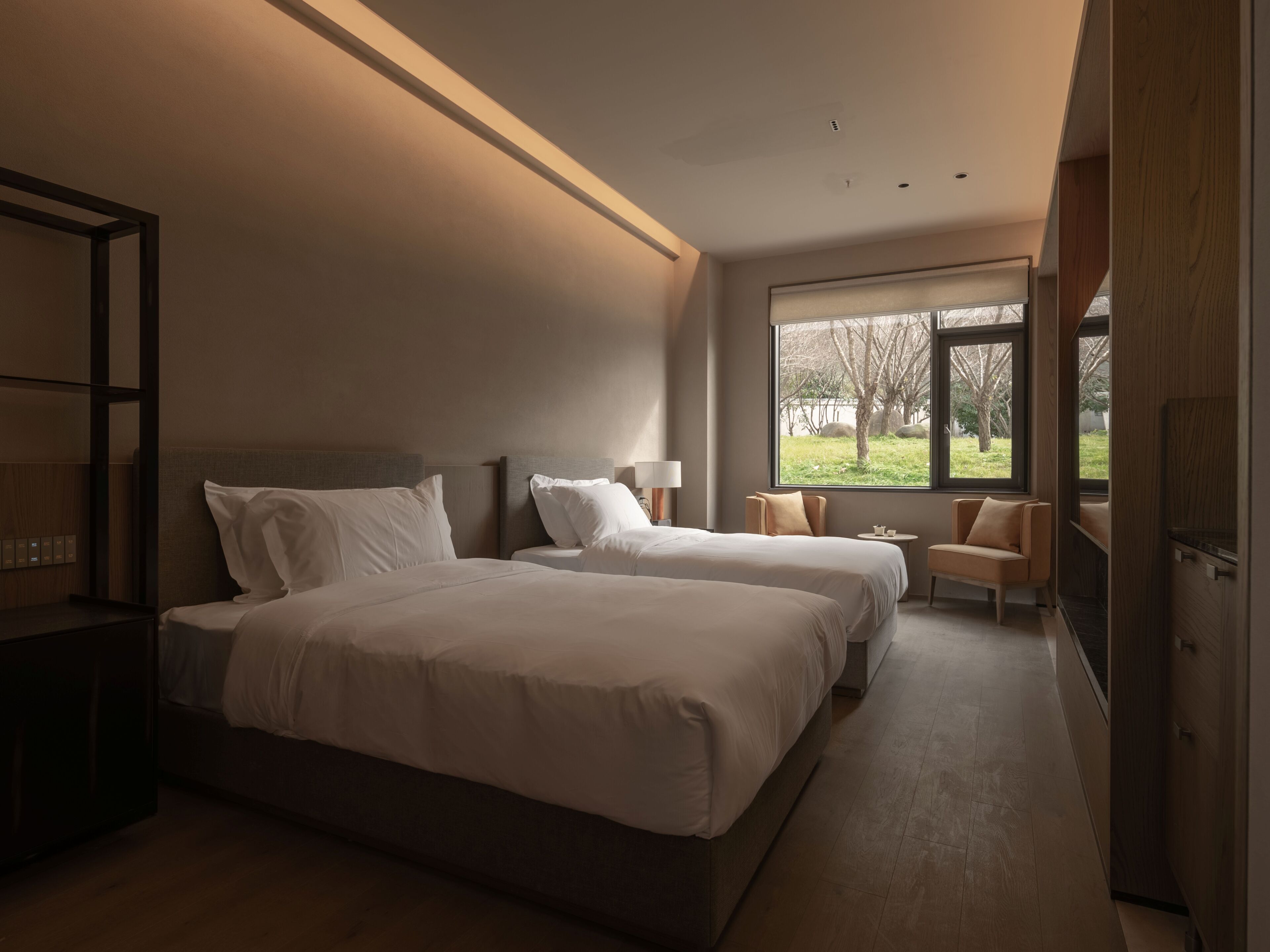 A modern hotel room with two beds, a sitting area, and a large window offering a view of the outdoors.
