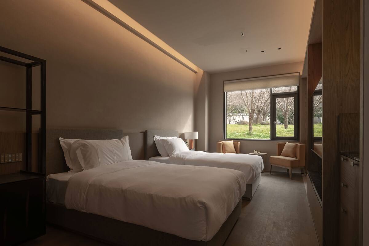 A modern hotel room with two beds, a sitting area, and a large window offering a view of the outdoors.