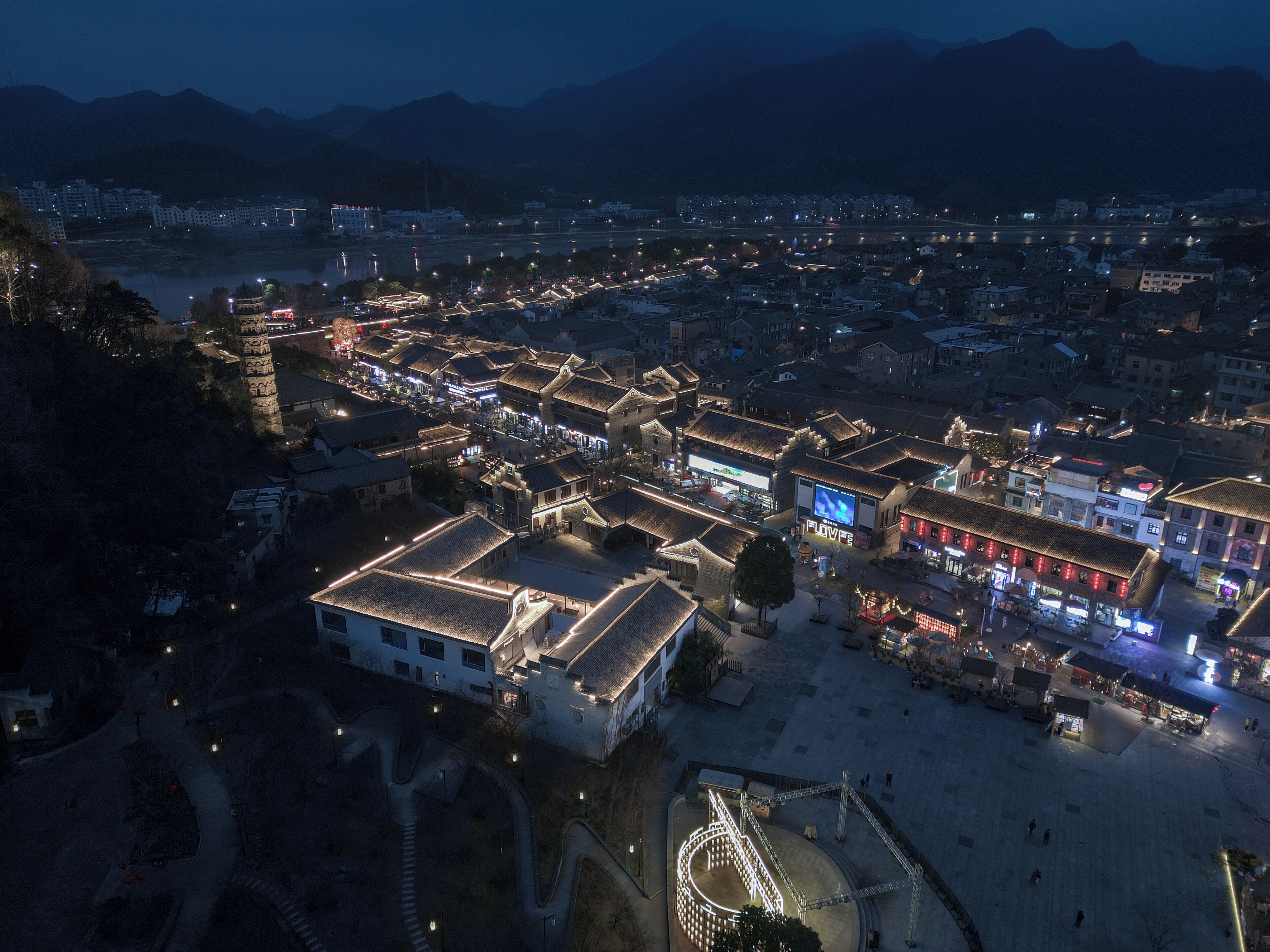 An aerial view of a town at night with illuminated buildings, a river, and mountains in the background.
