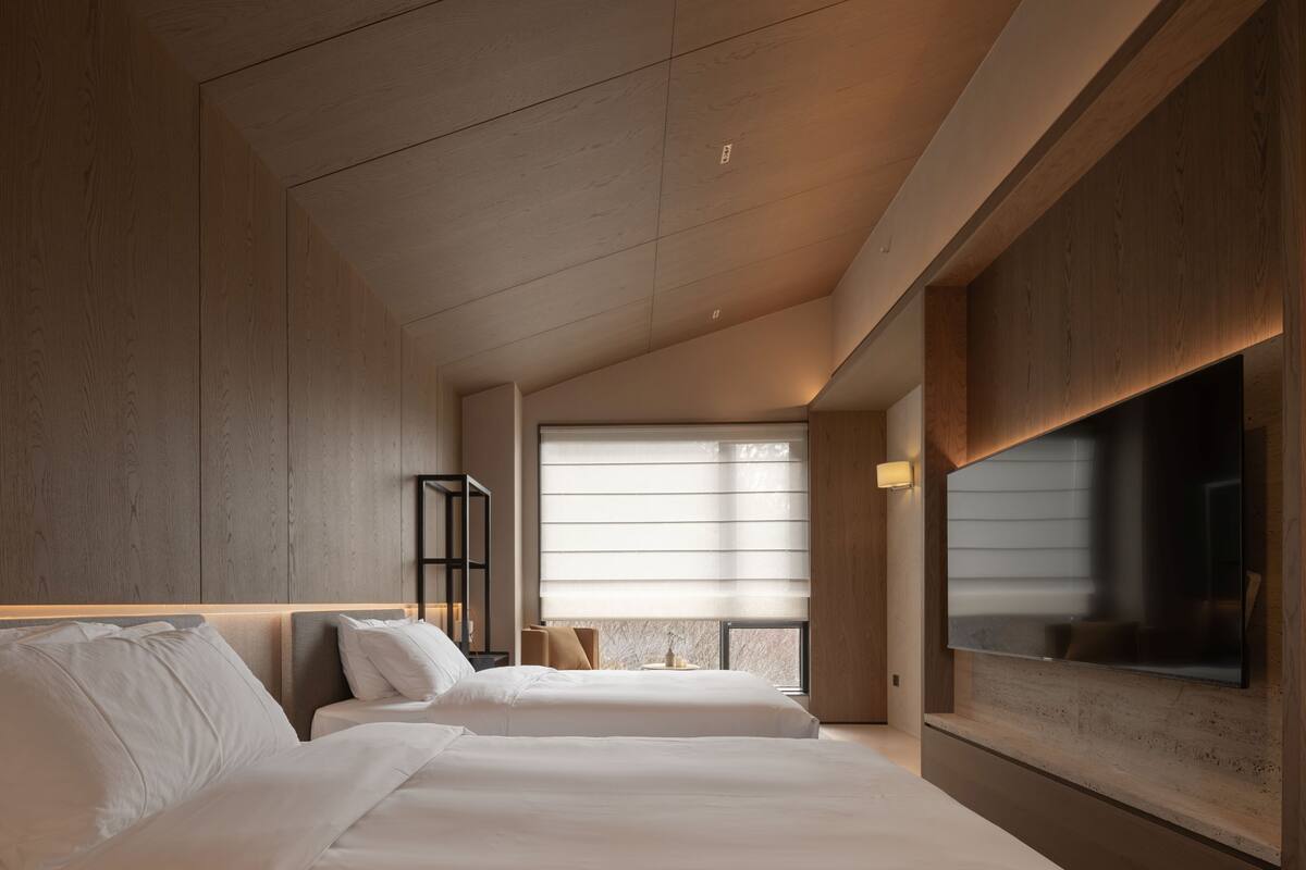 Una habitación de hotel moderna con dos camas, un ventanal con persianas, un televisor de pantalla plana montado en la pared y techo de madera.