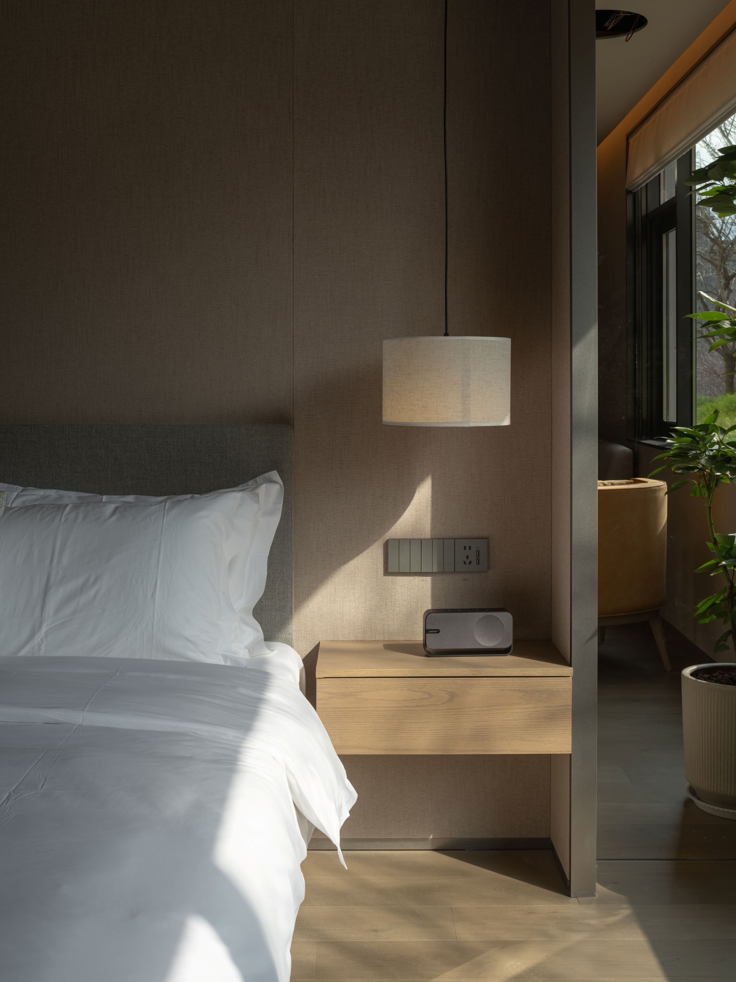 Una habitación de hotel moderna con cama, mesita de noche, lámpara, enchufe y una planta en maceta.