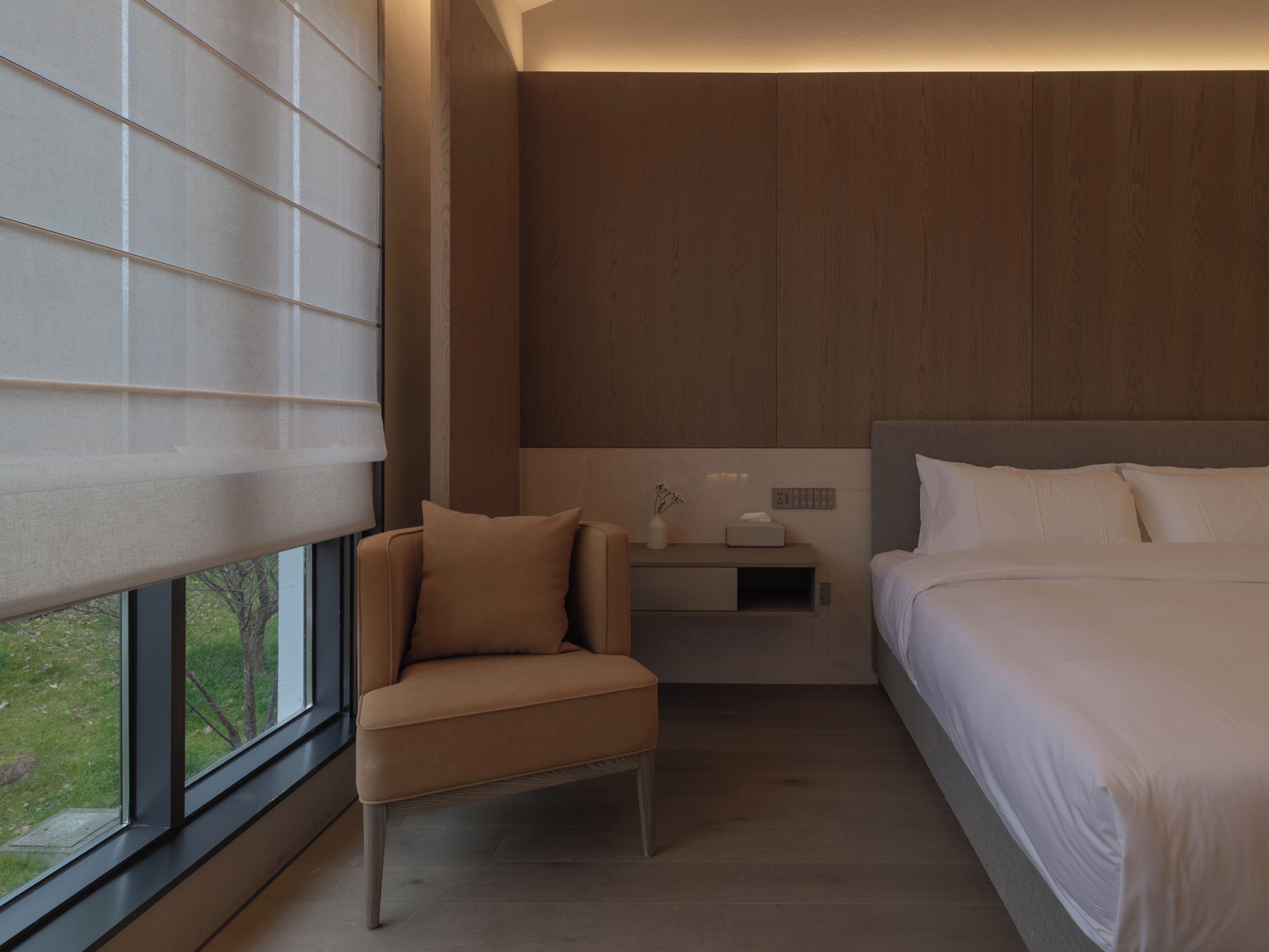 A modern hotel room with a bed, a chair, a nightstand, and a window with blinds.