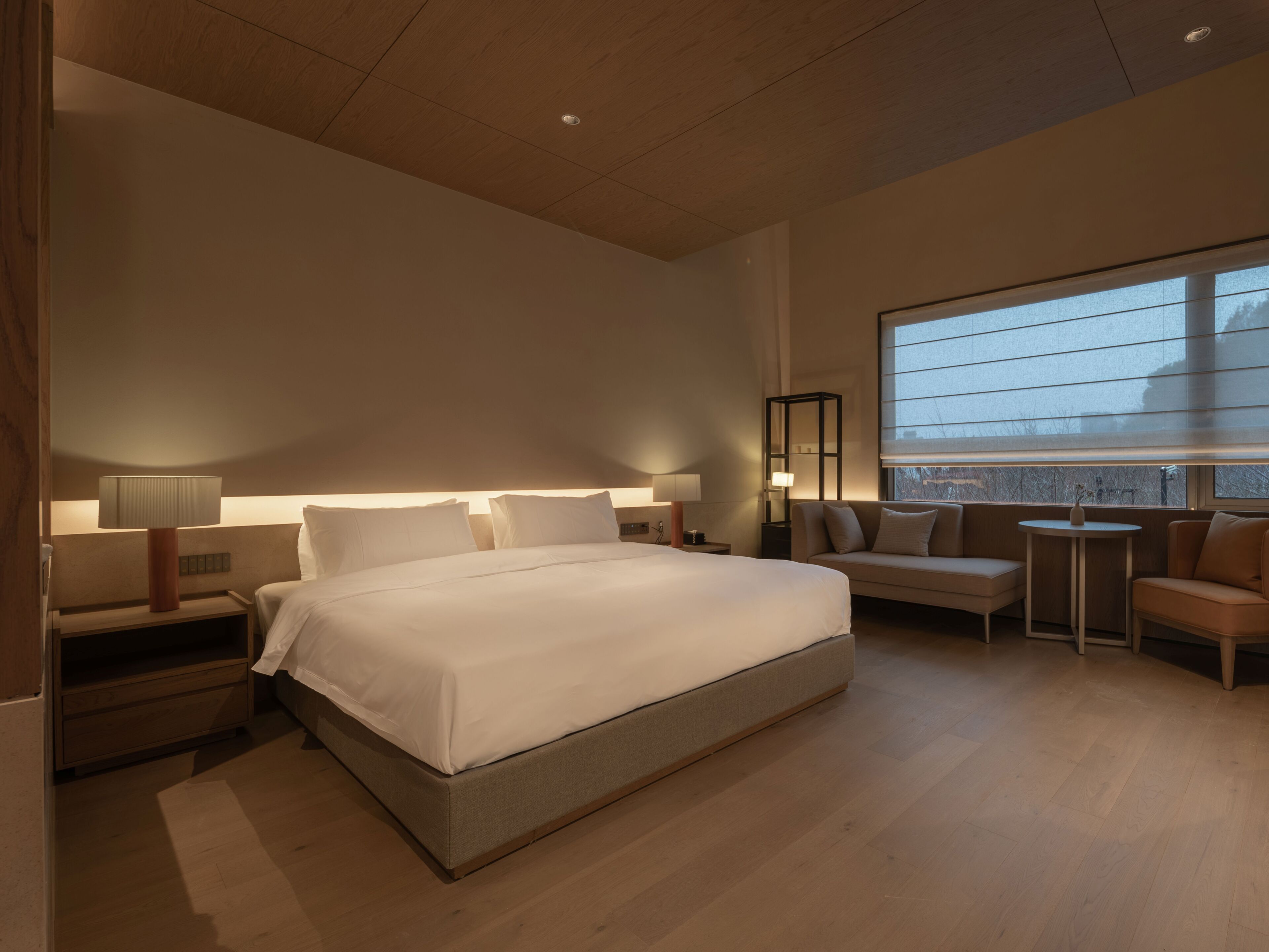 A modern hotel room with a large bed, bedside tables, a sofa, a small table, and a large window.