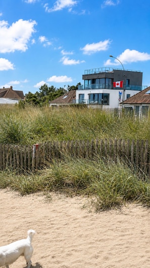 A modern building with a Canadian flag, surrounded by tall grass and a wooden fence. - Exceptional Seafront Apartment • 180° Sea View • Walk to Beach • D-Day Beaches (Colleville-Montgomery)