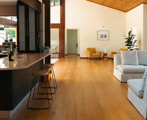 A spacious living area with a wooden floor, a white sofa, a yellow armchair, and a bar area with stools. - Spacious 5-bedroom house with mountain views (Cape Town)