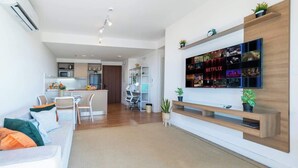 Una sala de estar moderna con un sofá, un televisor montado en la pared y una cocina al fondo.
