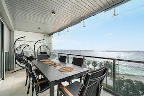 Un comedor con vista al océano, que incluye una mesa larga con individuales tejidos, sillas de alambre negro y una barandilla de vidrio con vista al mar.