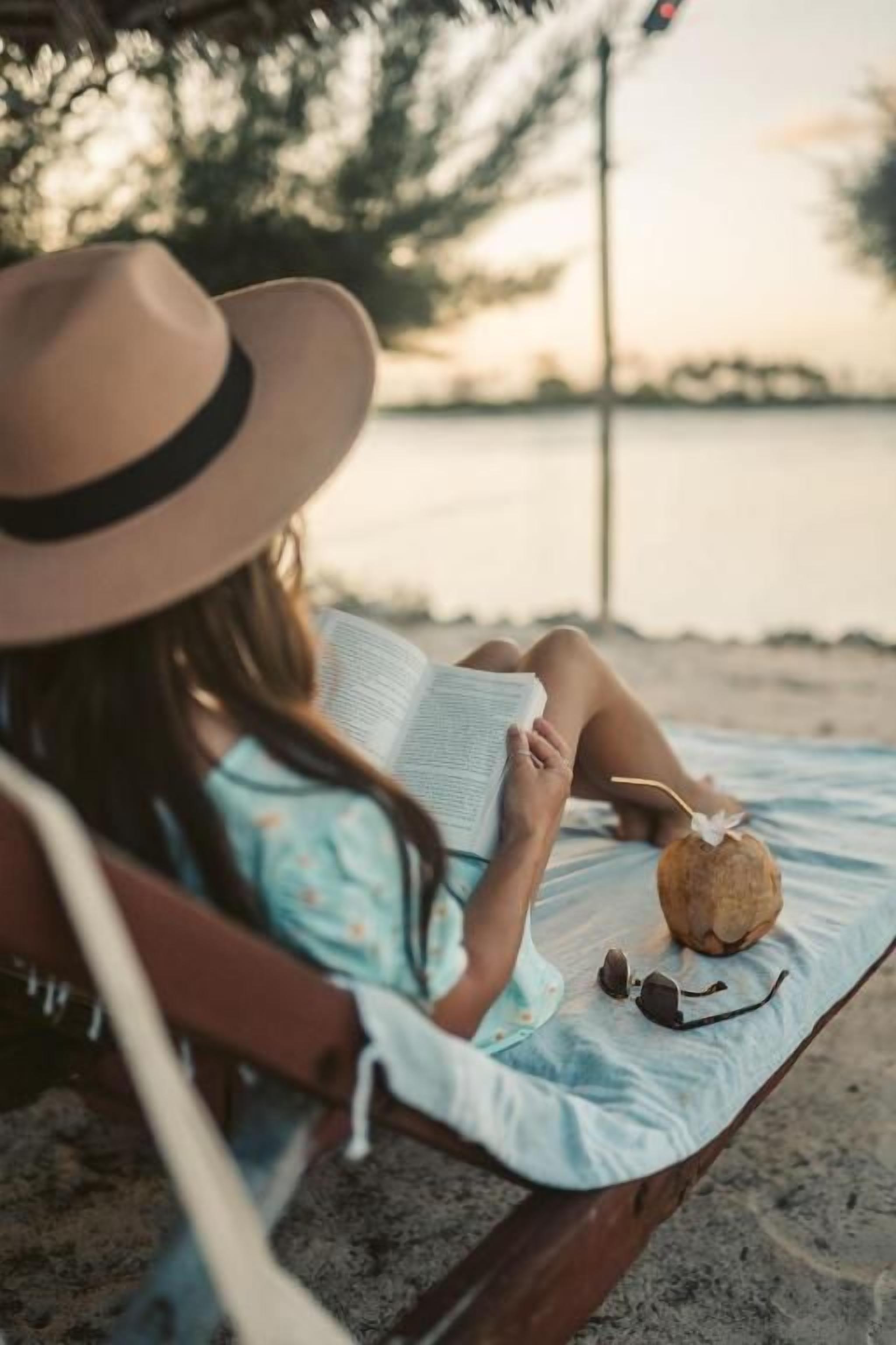 A person sitting on a beach chair reading a book with a straw hat and sunglasses beside them.