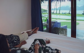 Een slaapkamer met een bed, een raam met gordijnen en uitzicht op een zwembad en groen.