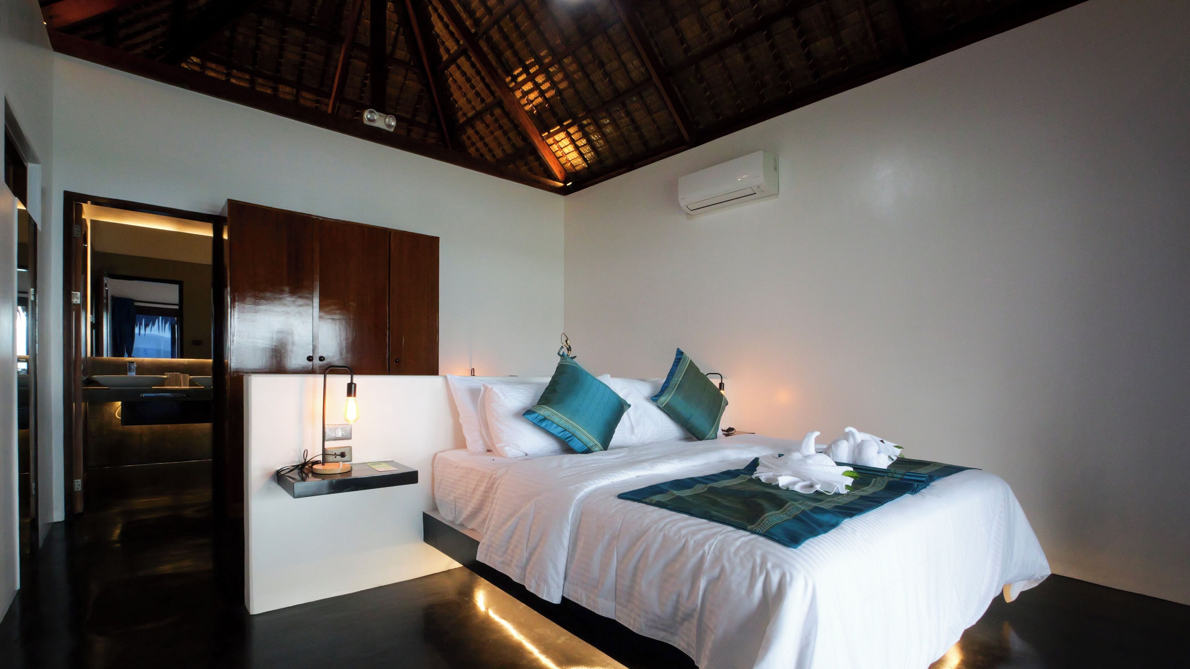 Un dormitorio con cama, almohadas, mesita de noche, lámpara, espejo y aire acondicionado.