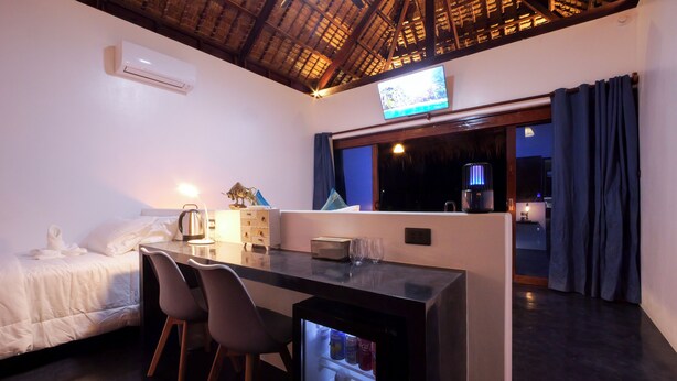 A hotel room with a bed, a desk with chairs, a TV, a minibar, and a window with curtains.