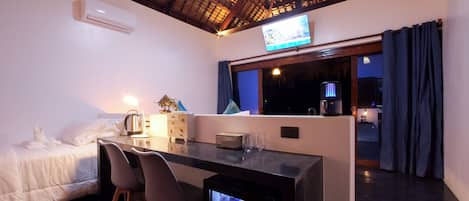 A hotel room with a bed, a desk with chairs, a TV, a minibar, and a window with curtains.