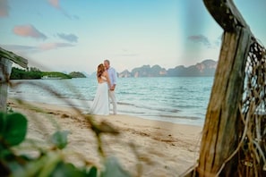 Una pareja de pie en la playa, con una estructura de madera al frente y un barco en la distancia.