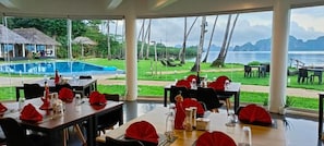 Un área de comedor al aire libre con mesas dispuestas para dos, con vista a una piscina y al agua.
