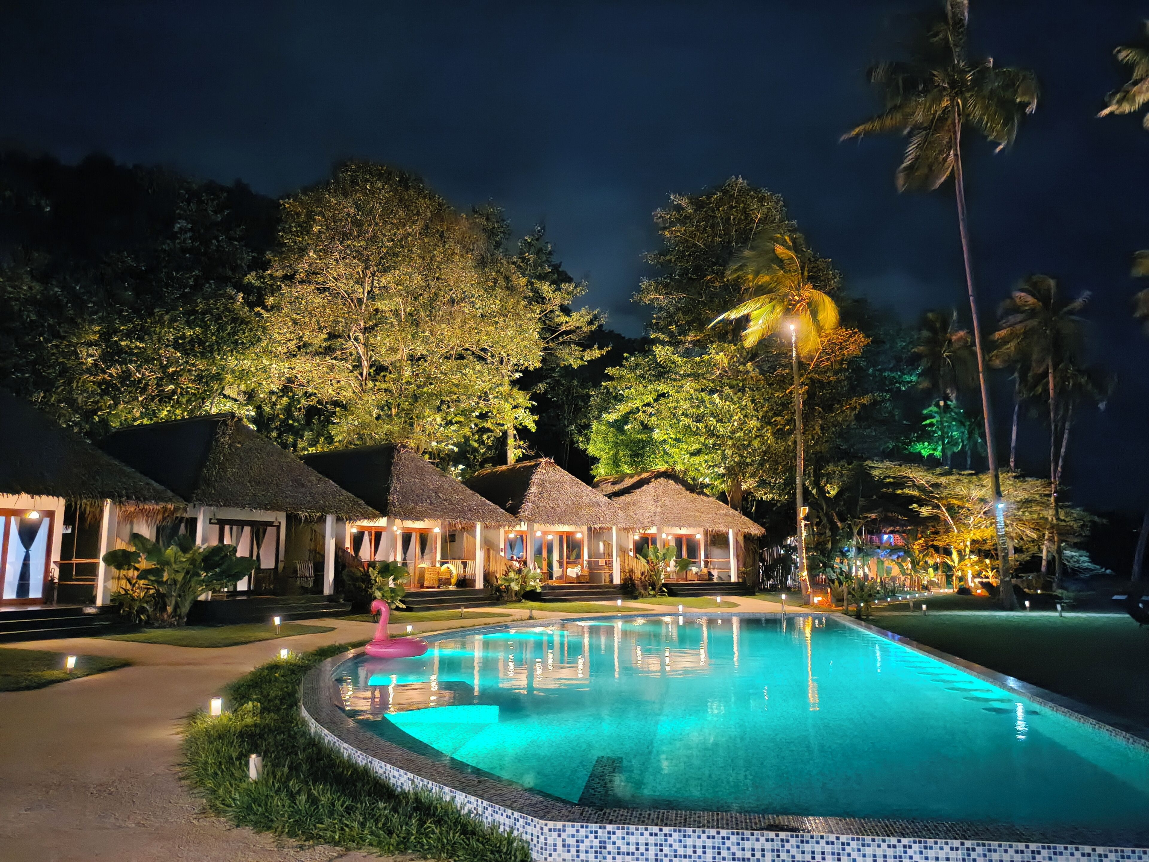 A nighttime resort with a swimming pool, thatched-roof huts, and tropical vegetation.