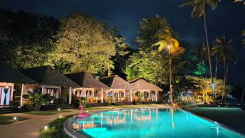 A nighttime resort with a swimming pool, thatched-roof huts, and tropical vegetation.