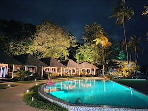 A nighttime resort with a swimming pool, thatched-roof huts, and tropical vegetation.