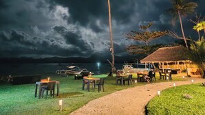 A beachside dining area with tables and chairs, a thatched-roof structure, and a palm tree.