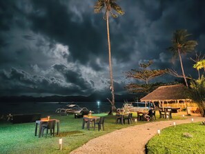 A beachside dining area with tables and chairs, a thatched-roof structure, and a palm tree. - Bebeladan Beach Resort (El Nido)