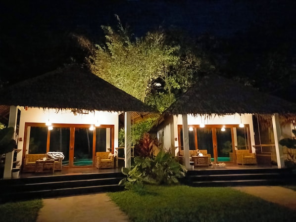 Two thatched-roof structures with outdoor seating, a pathway leading to them, and a large tree in the background.