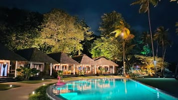 Un resort nocturno con piscina, construcciones de techo de paja y vegetación tropical.