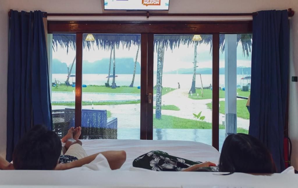 A room with a bed, two people lying down, and a large window with a view of a beach and palm trees.